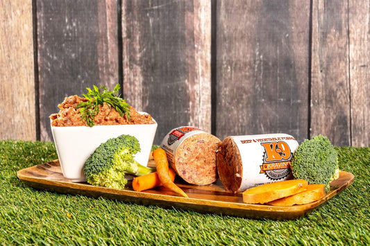 Raw dog food displayed with fresh vegetables including broccoli, carrots, and sweet potato on a wooden platter.