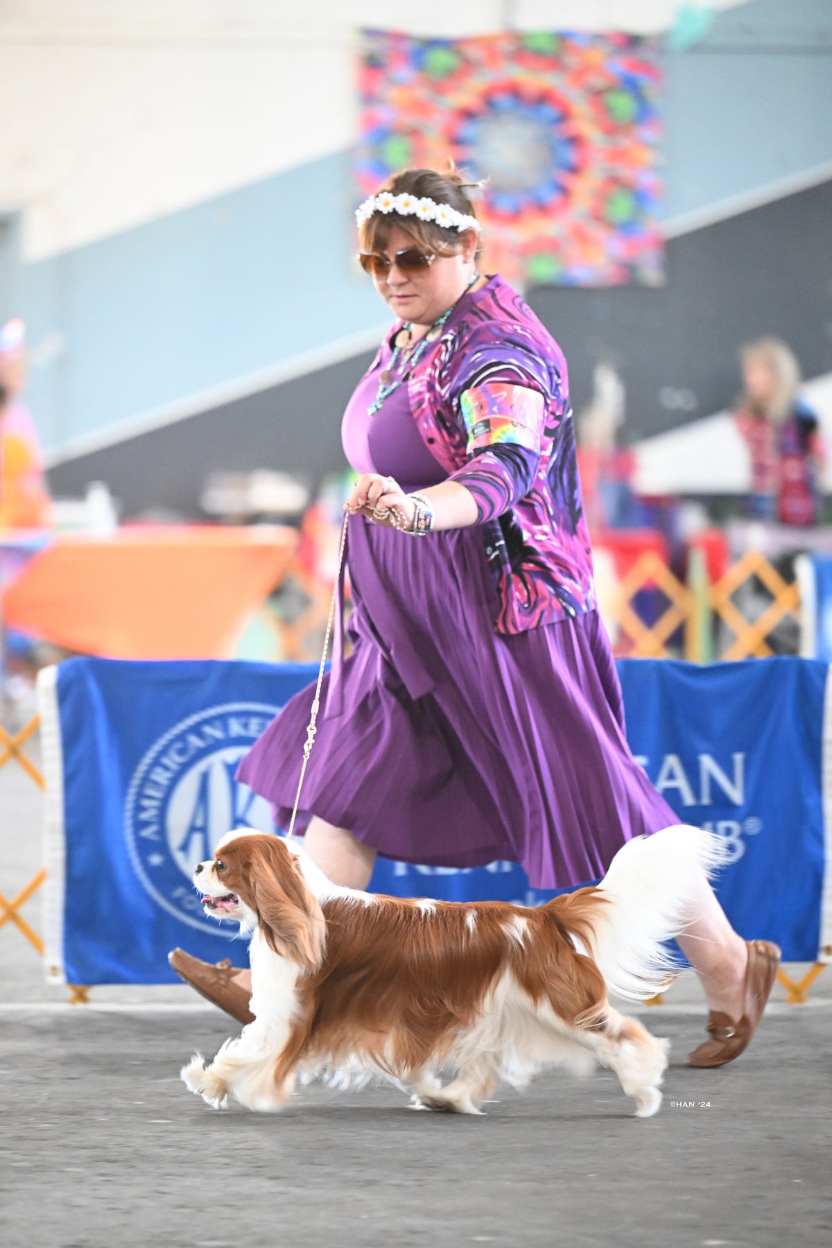 Handler in a purple dress and flower crown leads a Blenheim Cavalier King Charles Spaniel at a dog show.