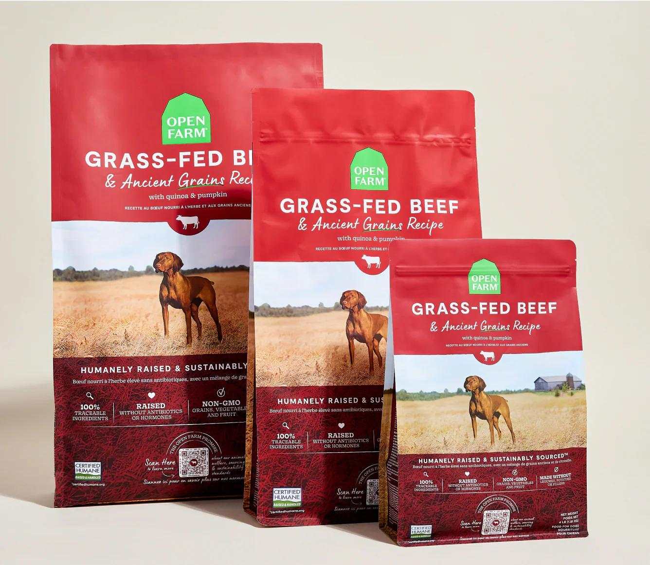 Three sizes of Open Farm Grass-Fed Beef & Ancient Grains Recipe dog kibble bags with a brown dog on a field background