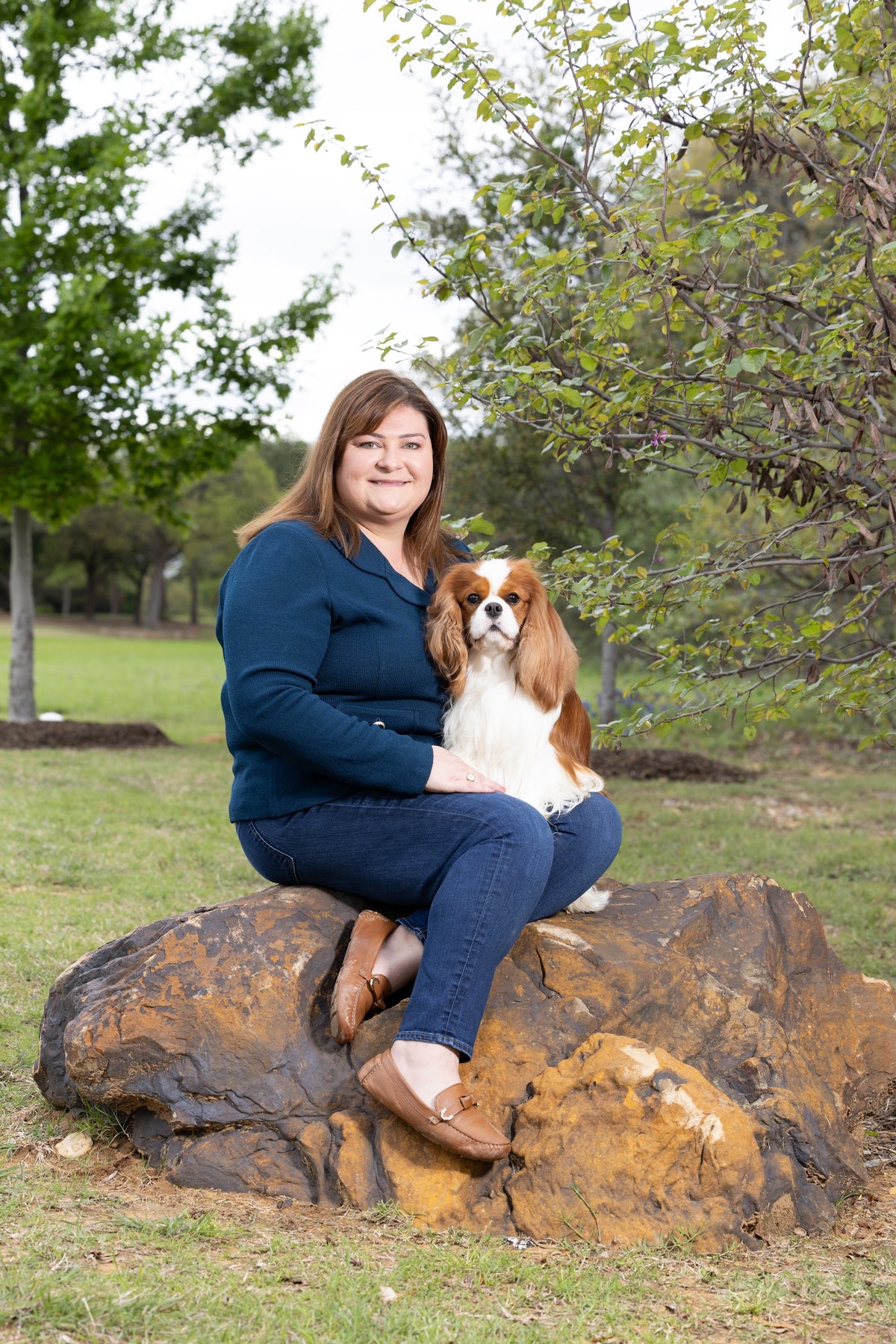 Woman in a blue blazer and jeans sitting on a large rock outdoors with her Cavalier King Charles Spaniel.
