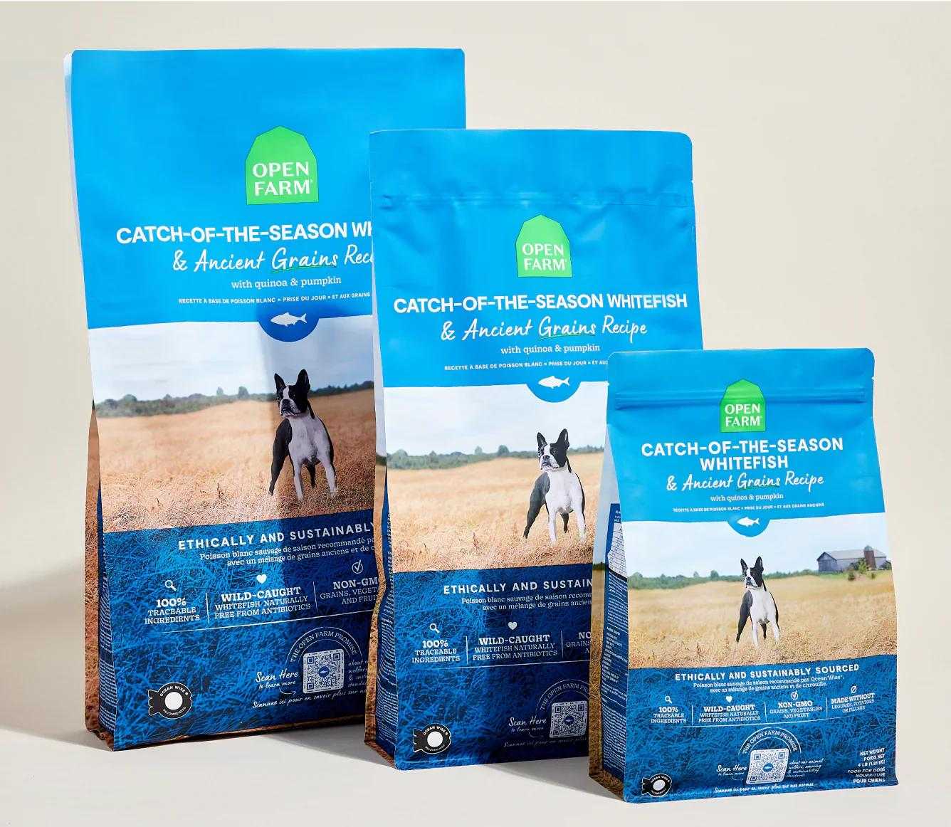 Three blue bags of Open Farm Catch-of-the-Season Whitefish & Ancient Grain Recipe dog kibble with a dog image and natural field background