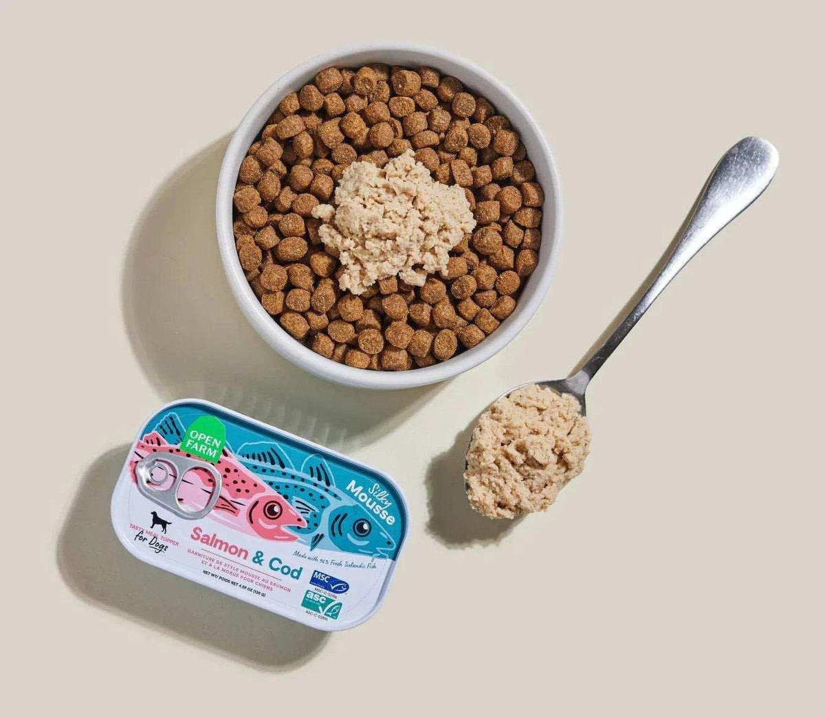 Salmon & Cod Topper in a blue-and-pink rectangular tin, with a beige mousse texture shown on kibble and a spoon.